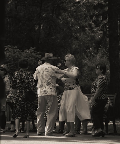 A group of elderly people is gathered in a park, engaging in social interaction. Some are dancing while others watch and converse. The setting is shaded by trees, creating a peaceful and intimate atmosphere.