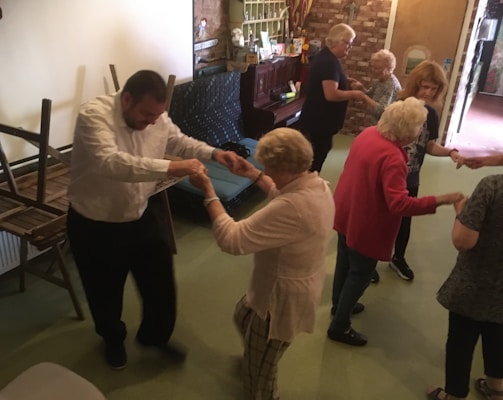 A group of elderly people are engaging in a dance activity in a room. Some chairs are stacked on the side, and a piano is visible in the background. The atmosphere appears lively and social.